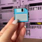 Retro Floppy Disk Enamel Pin - A nostalgic enamel pin featuring a vintage floppy disk with a smiling face, perfect for tech enthusiasts and lovers of retro accessories.