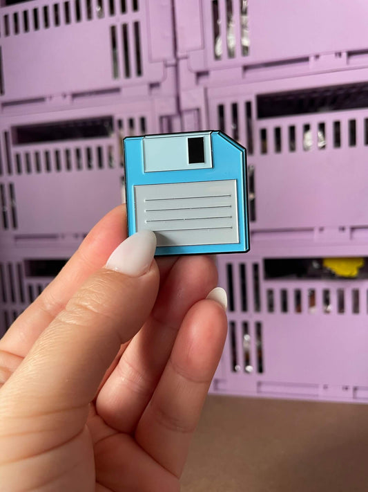 Retro Floppy Disk Enamel Pin - A nostalgic enamel pin featuring a vintage floppy disk with a smiling face, perfect for tech enthusiasts and lovers of retro accessories.